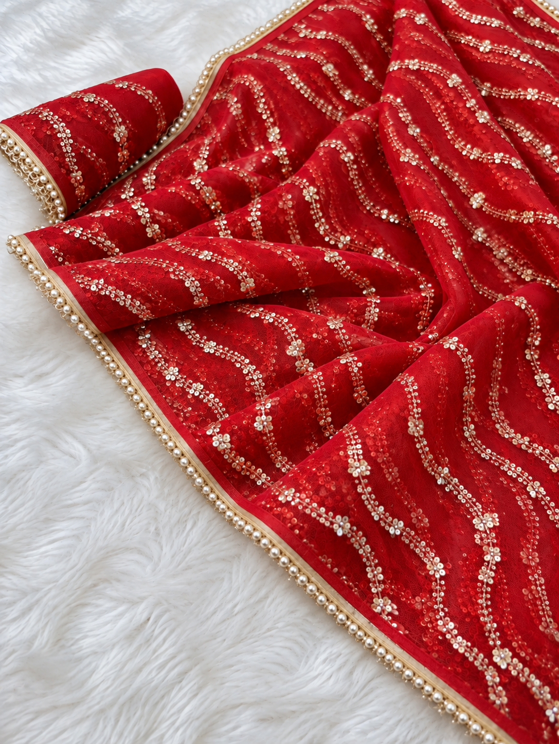 Red Net Saree Pearl Sequin Embroidered (Karwa Chauth Saree) A red fabric accessory, likely a saree drape, decorated with gold sequins in wavy stripes and finished with a gold beaded border, laid out on a white faux fur surface.