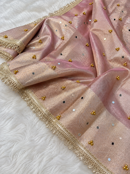 A ghungru saree draped pink organza fabric with gold zari embroidery, gold and silver mirror work, and a decorative gold border, laid flat on a white faux fur surface.