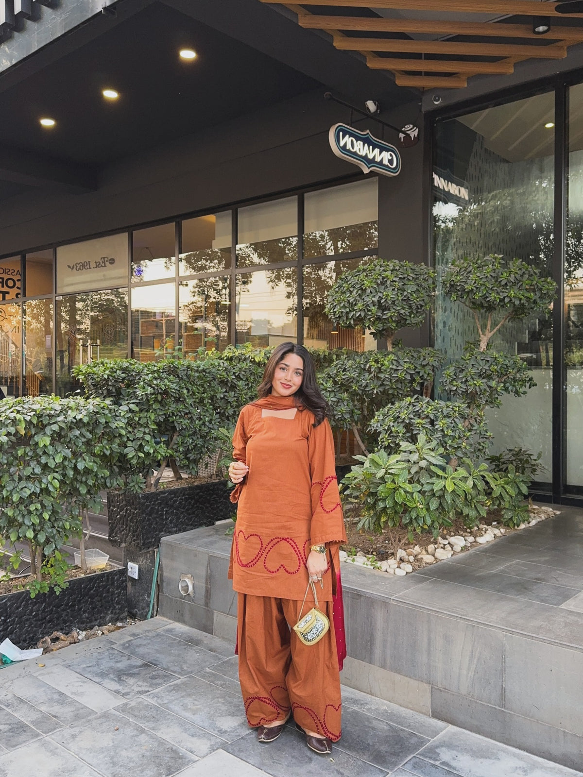 A woman stands outdoors in front of a modern building entrance wearing a rust-orange traditional outfit with matching wide-leg pants and a red draped scarf. The background features green plants, glass windows, and a dark facade.Designer Brown Farshi suit for women