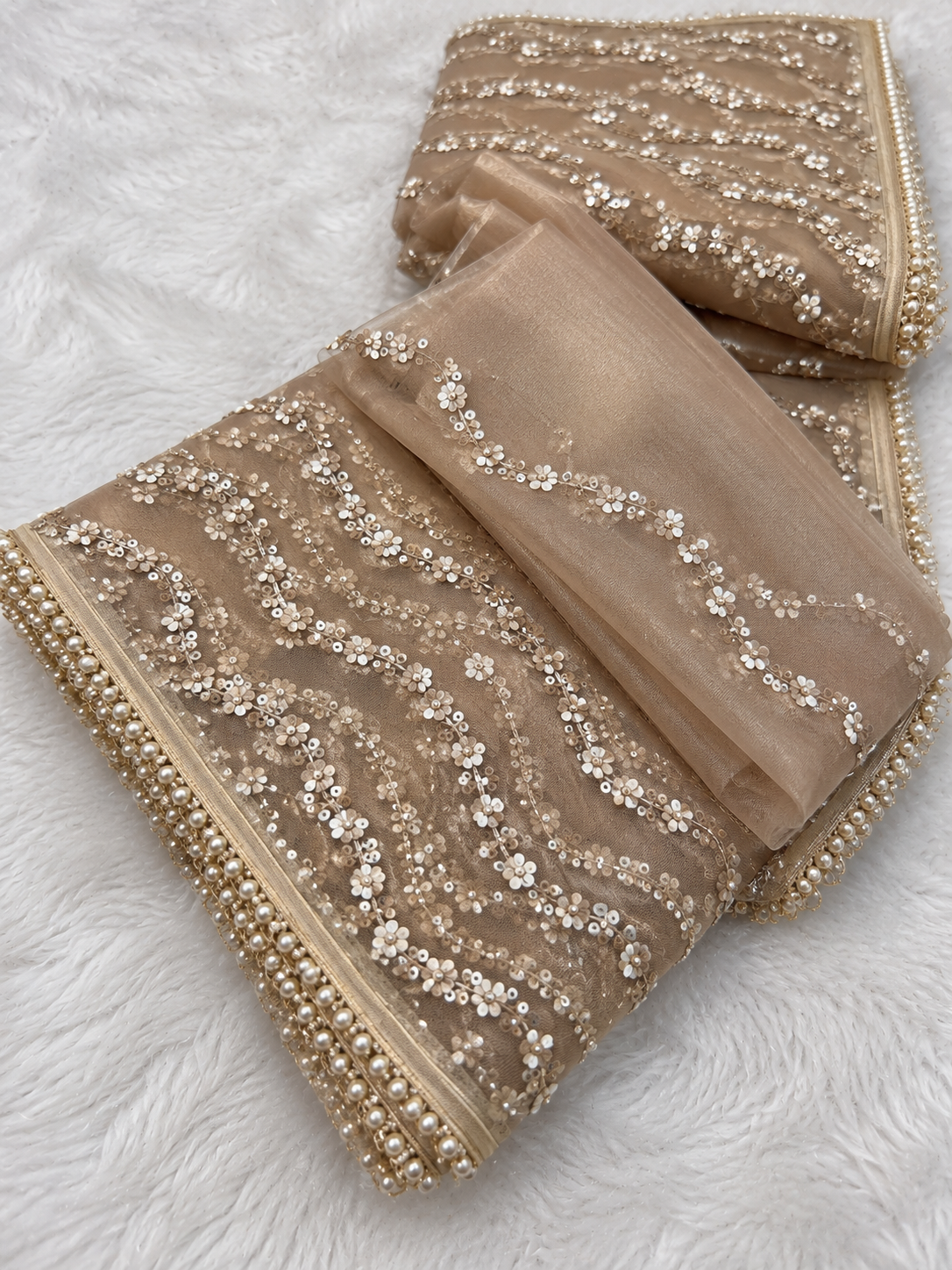 Golden Net Saree for Women, Pearl and Sequin Embroidered Designer Saree .folded organza fabric set with intricate silver and gold floral embroidery and bead borders, displayed on a white faux fur background.
