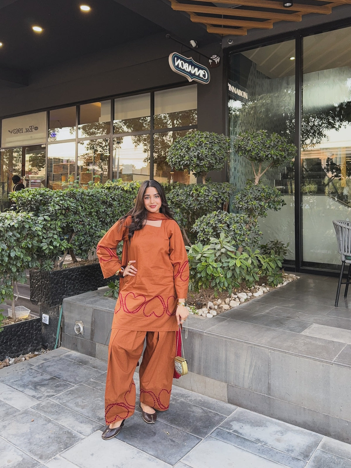 A woman stands outdoors in front of a modern storefront wearing a matching rust-orange two-piece outfit with red embroidered heart patterns. The set includes a long-sleeve top and wide-leg pants, paired with dark pointed shoes and a small pink handbag, Designer Farshi suit for women