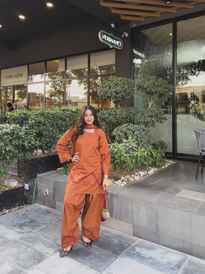 A woman stands outdoors in front of a modern storefront wearing a matching rust-orange two-piece outfit with red embroidered heart patterns. The set includes a long-sleeve top and wide-leg pants, paired with dark pointed shoes and a small pink handbag, Designer Farshi suit for women