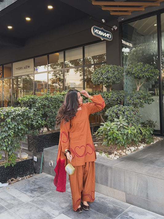 A woman wearing a rust-orange embroidered  Farshi kurta and matching wide-leg Farshi pants stands outside a modern shop with greenery. She holds a red shawl and a small patterned clutch.