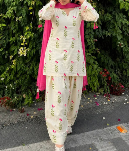Person wearing a traditional outfit Farshi Suit with floral patterns in front of green foliage.