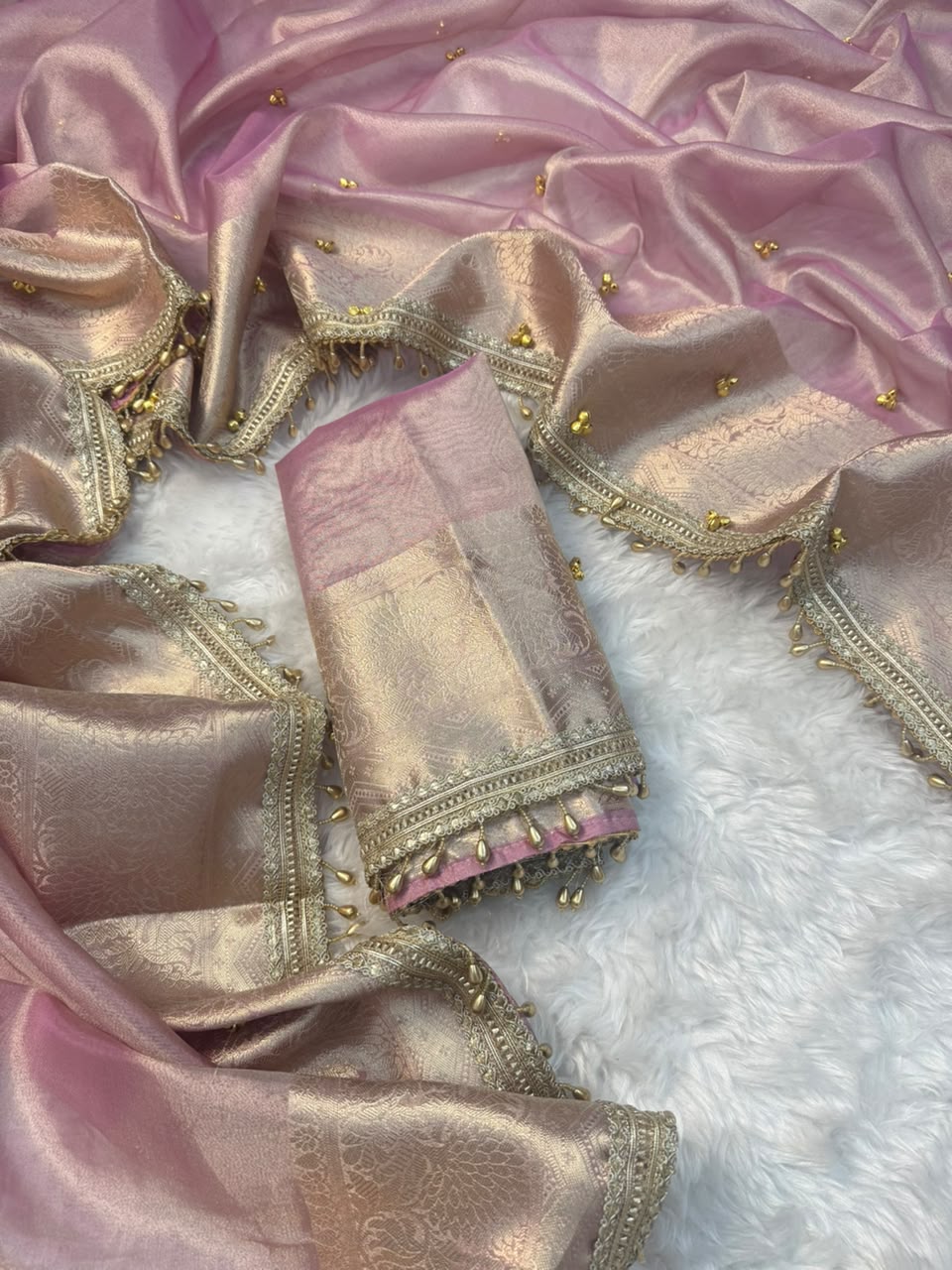 Ghungroo saree A folded pink and gold zari fabric textile with ornate metallic borders and small gold embellishments, arranged on a white faux-fur surface.