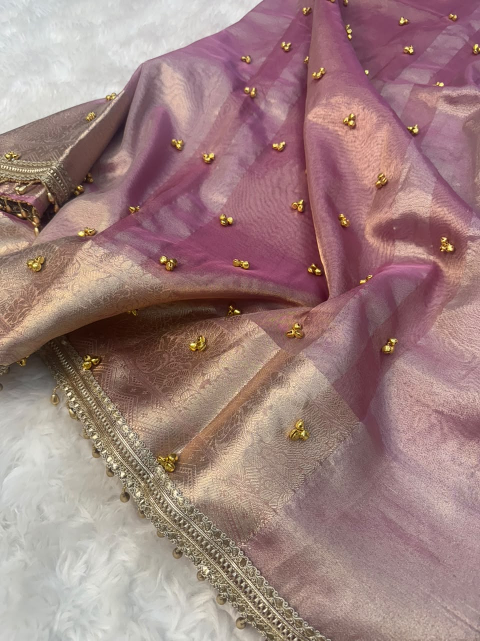 A close-up of a draped silk saree or fabric in a dusty mauve-pink and champagne-gold colorway, decorated with small gold ghungroo embroidered motifs and a detailed gold zari border.