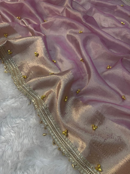 A close-up of a draped lavender-purple silk fabric with a shimmering metallic gold border, decorated with small gold floral motifs, laid over a white faux fur surface.