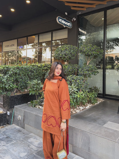 A woman wearing a rust-orange two-piece kurta set with red embroidered heart patterns stands in front of a modern building with green hedges and a 'MAMMO' sign. Trending Farshi suit
