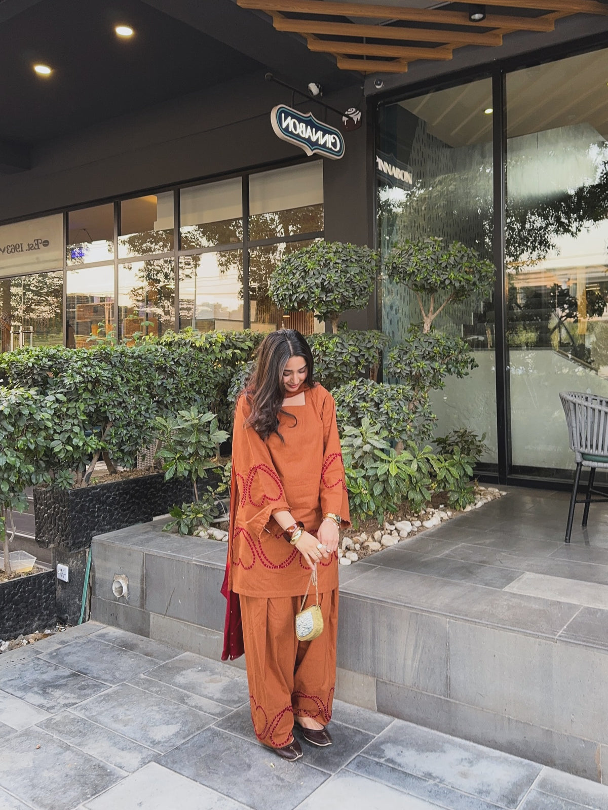 A woman wearing a rust-orange two-piece kurta set with red embroidery stands outside a modern building with glass windows and greenery, Rust brown Farshi suit salwar