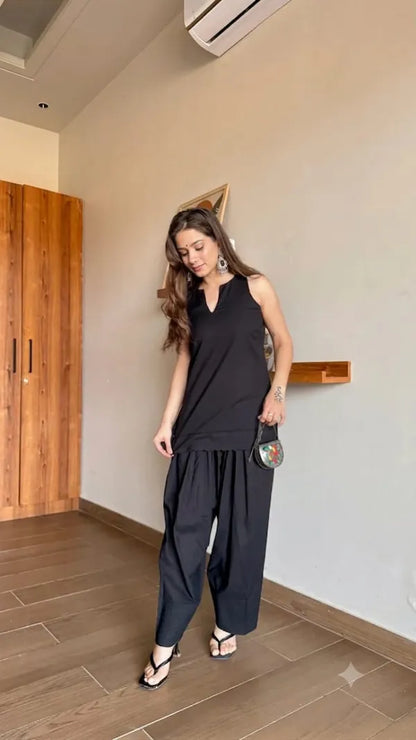 Woman in a black Farshi Suit outfit standing in a room with wooden furniture and a light-colored wall.