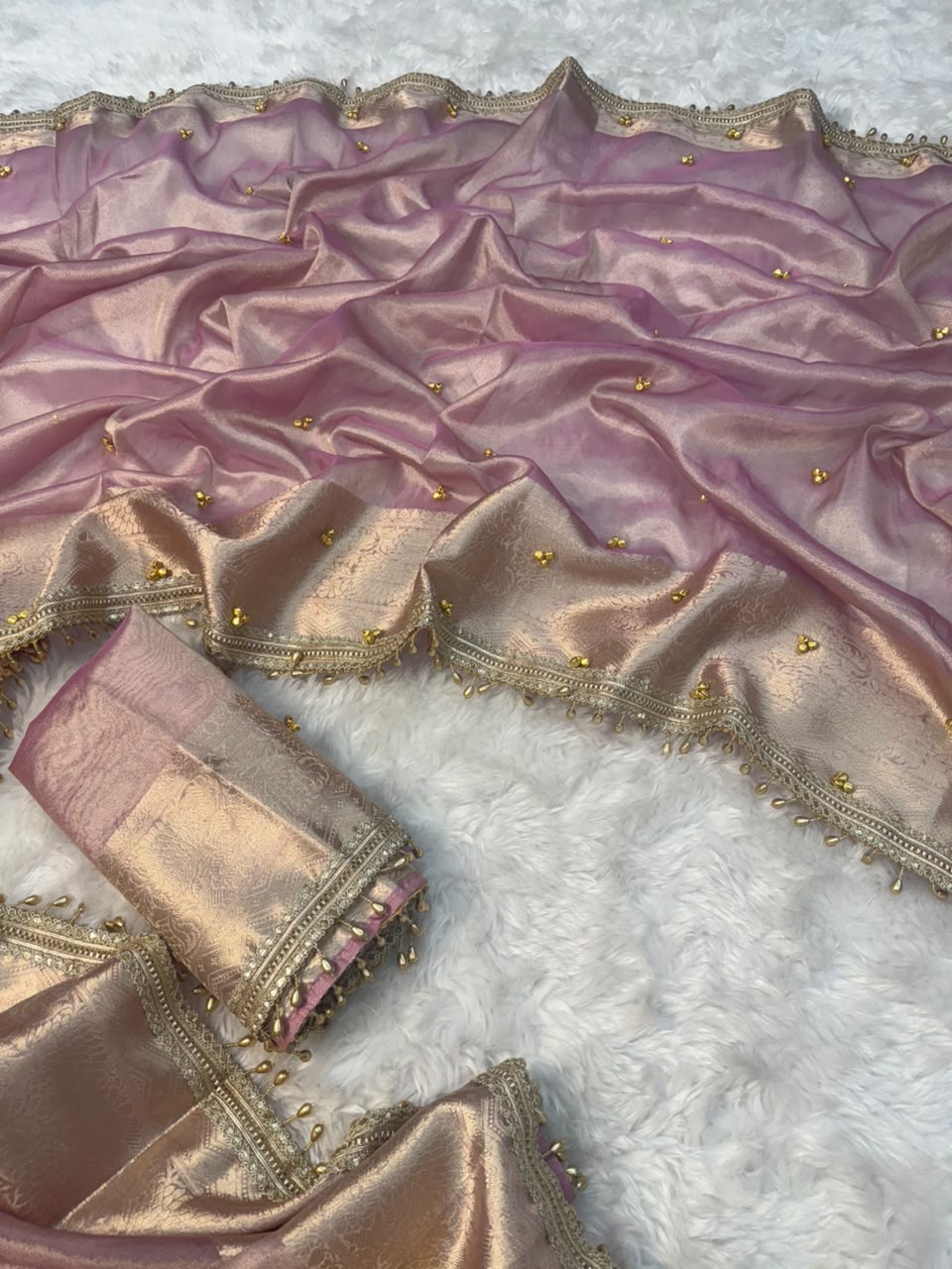 A draped pink and gold silk saree or fabric set with metallic zari borders, gold Ghungroo bead embellishments, and matching rolled fabric pieces, laid out on a white textured faux-fur surface.