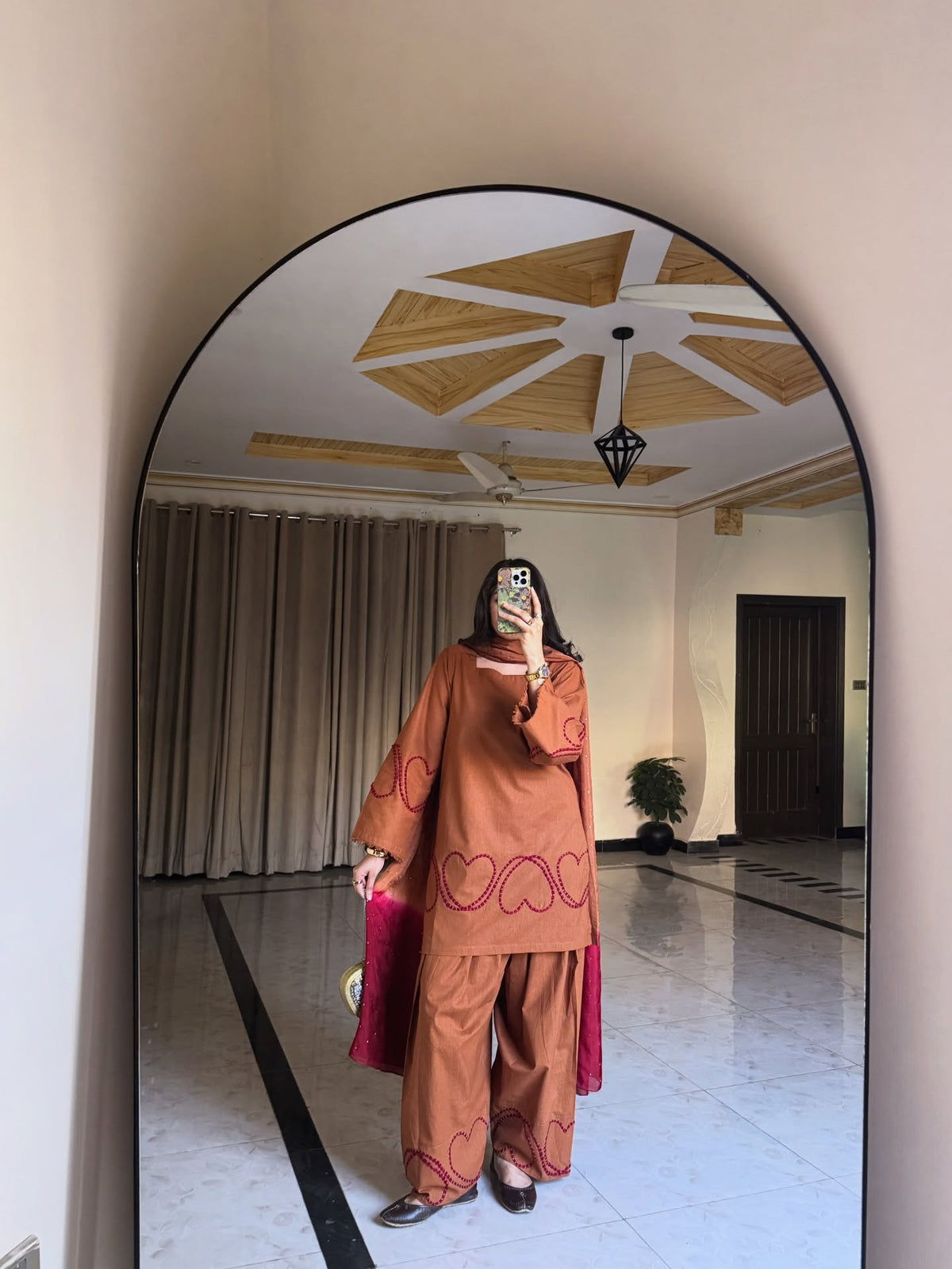A person takes a mirror selfie in a large indoor room, wearing a rust-orange two-piece outfit with embroidered heart patterns and a pink draped scarf or shawl, Rust brown Farshi suit salwar, 