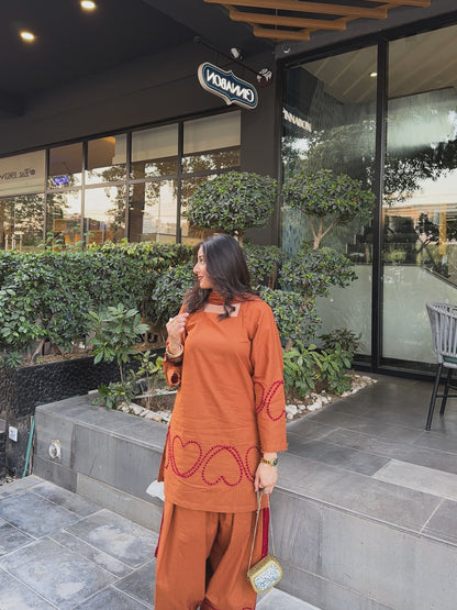 A woman wearing a rust-orange kurta and matching trousers with red heart embroidery, standing outside a modern building with greenery and a pool visible in the background., rust-brown-farshi-suit-salwar