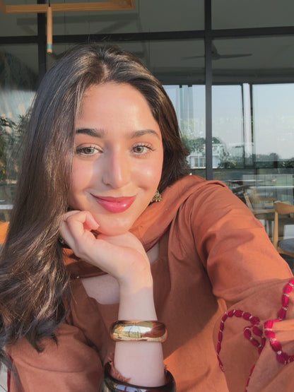 A woman with long dark hair resting her chin on her hand, wearing a rust-orange top and a gold-toned bangle, photographed in a bright indoor setting with large windows. rust-brown-farshi-suit-salwar, Heart-pattern decorative embroidery, Festive wear suit for women