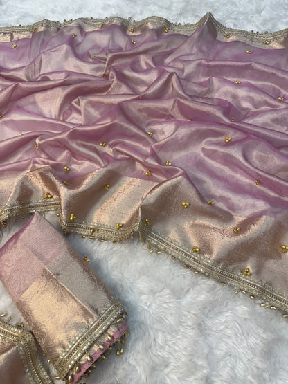 A draped pink and gold silk saree or fabric piece with metallic zari borders, gold ghungroo bead embellishments, and a matching rolled fabric piece on a white faux fur background.